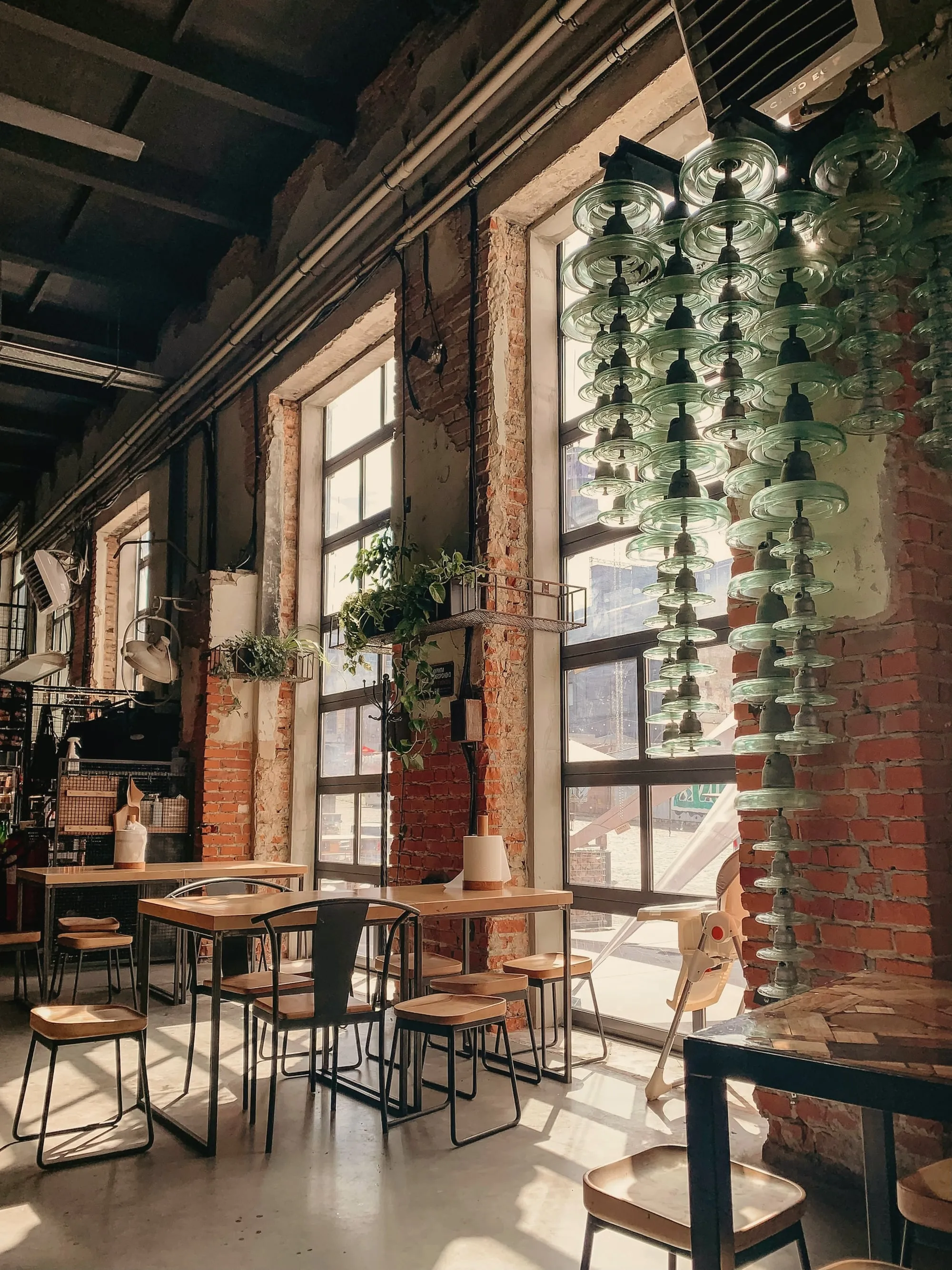 Café tables under tall steel-framed windows, with brick walls and hanging plants